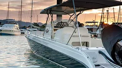 White motorboat with dark canopy docked in marina at sunset; sailboats in background.