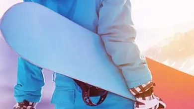 A person in a blue snowsuit holds a snowboard under one arm, standing in front of snowy mountains with the sun shining overhead.