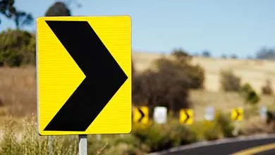 A yellow road sign with a black right-pointing chevron warns of a sharp right curve; additional road signs are visible further ahead.