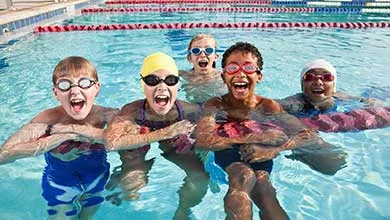 Five children in swimsuits and goggles laugh and play together in a swimming pool.