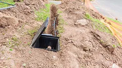 A narrow trench cuts through a dirt mound beside a road, containing an open black utility box with exposed cables.