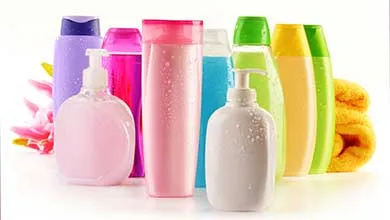 Colorful plastic shampoo and soap bottles on a white surface, with a yellow towel and pink flowers in the background.